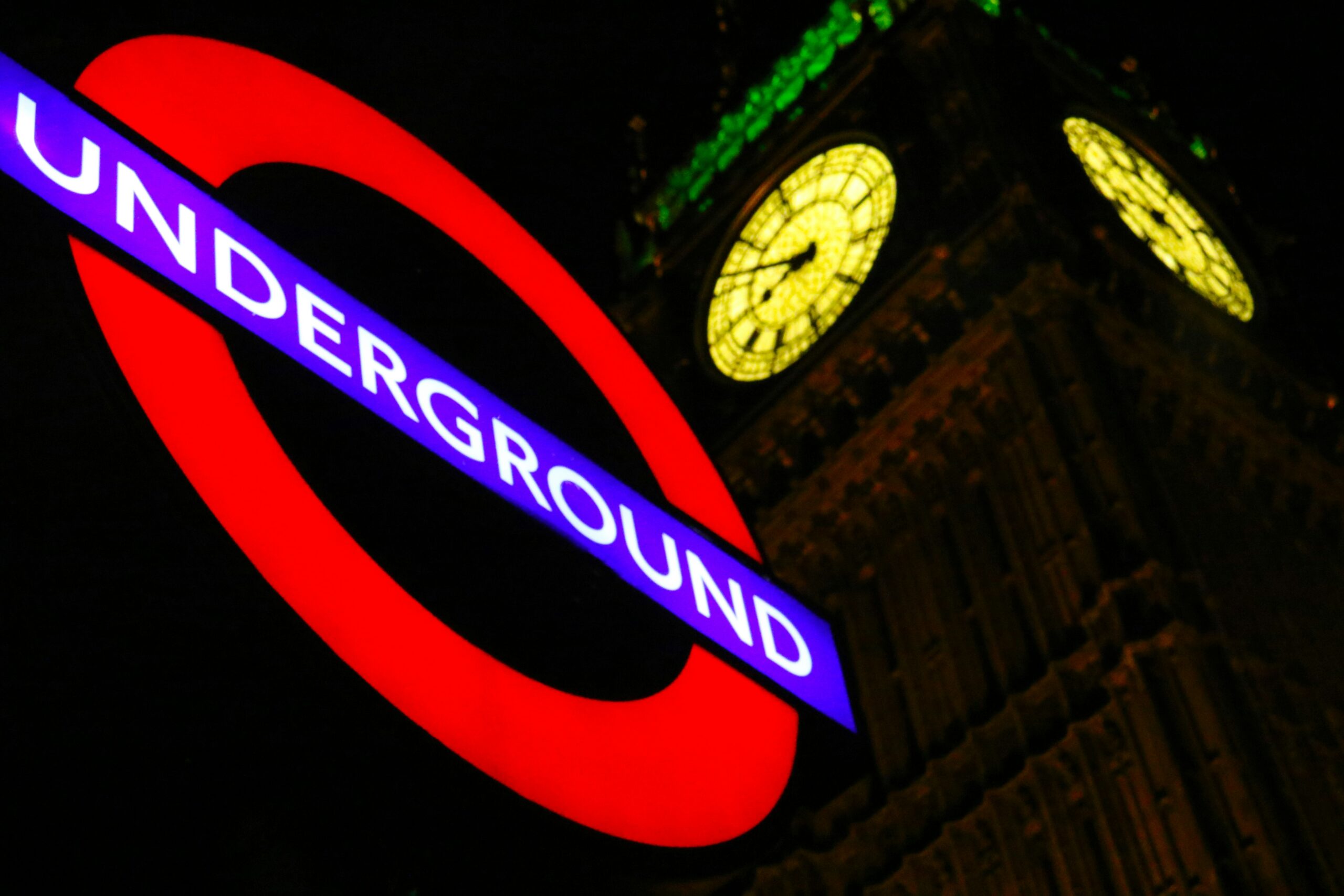 Lit up Underground sign with Big Ben in the background at night.